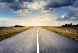 A long straight road stretches through fields under a dramatic cloudy sky.