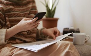 Crop unrecognizable female with newspaper text messaging on cellphone while sitting in armchair with cup of hot drink in house